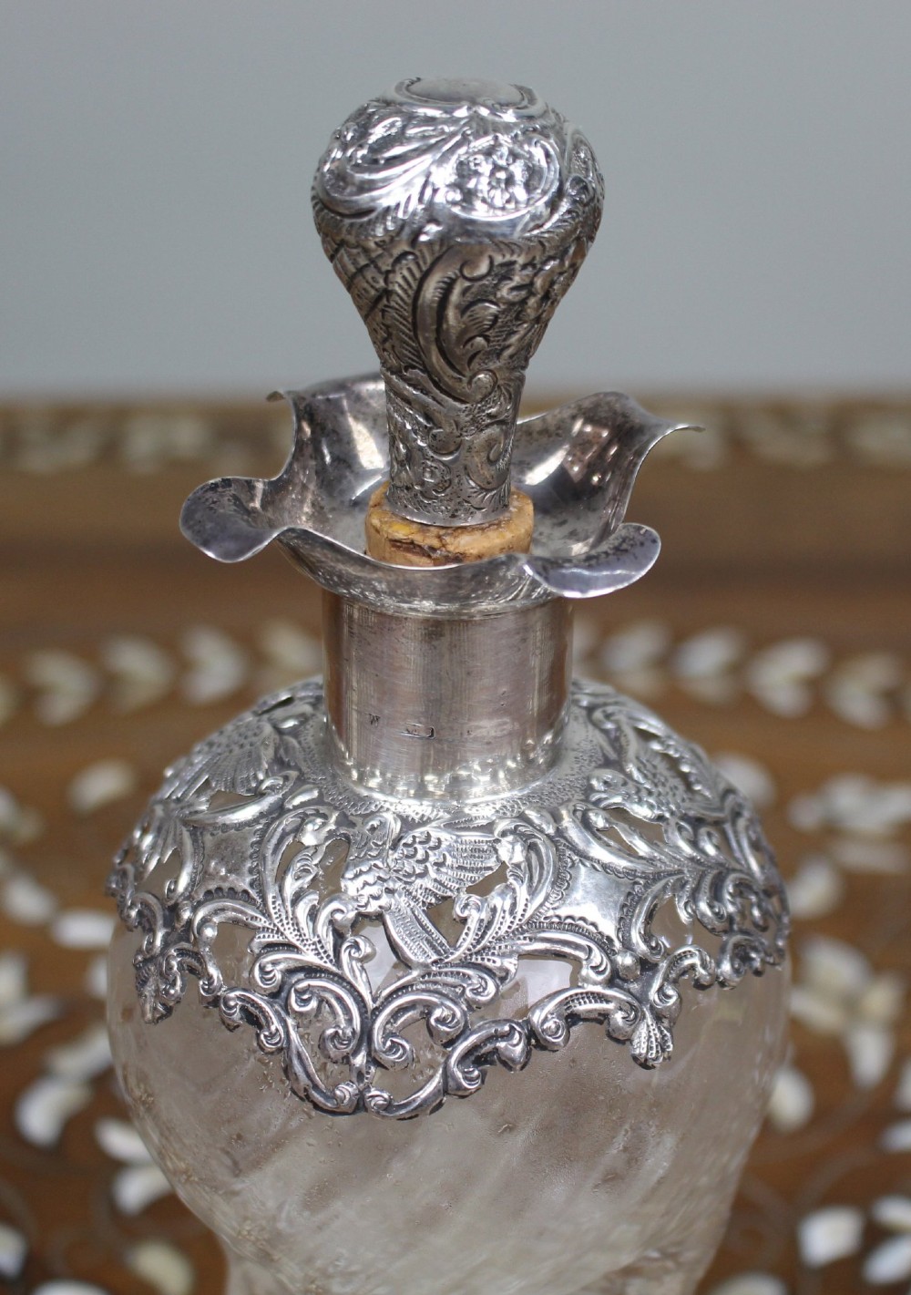 An antique clear glass decanter featuring a pinched waist, swirled body, ornate pierced silver shoulder mount, silver collar, and a chased silver stopper. View V