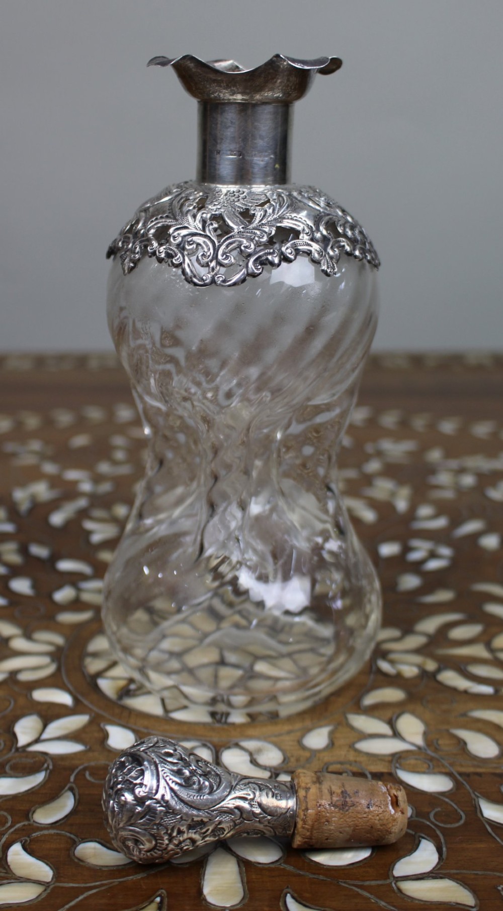 An antique clear glass decanter featuring a pinched waist, swirled body, ornate pierced silver shoulder mount, silver collar, and a chased silver stopper. View VI