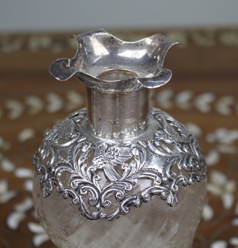 An antique clear glass decanter featuring a pinched waist, swirled body, ornate pierced silver shoulder mount, silver collar, and a chased silver stopper. View X