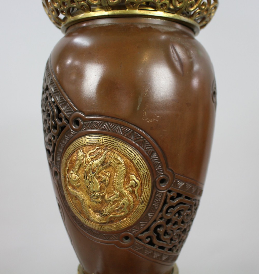 A Meiji period Japanese dark brown bronze oil lamp featuring reticulated openwork, a gilded dragon roundel, brass burner fittings, and decorative scroll feet. View X
