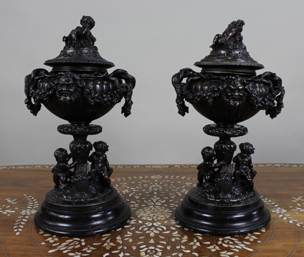 A pair of dark bronzed spelter urns with lids topped by seated putti figures, featuring Bacchic masks, relief garlands, and bases supported by sculpted cherubs.