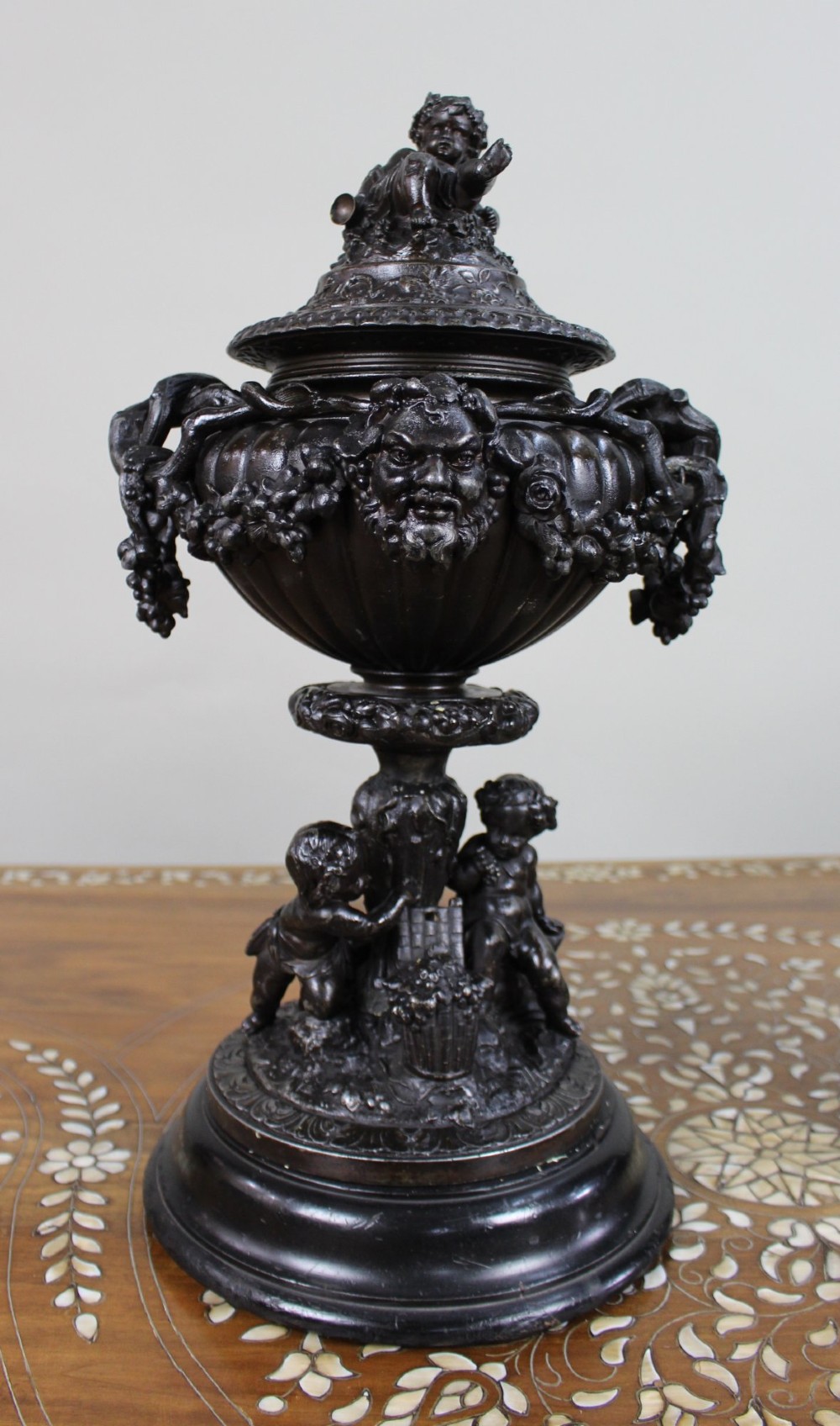 A pair of dark bronzed spelter urns with lids topped by seated putti figures, featuring Bacchic masks, relief garlands, and bases supported by sculpted cherubs. View II