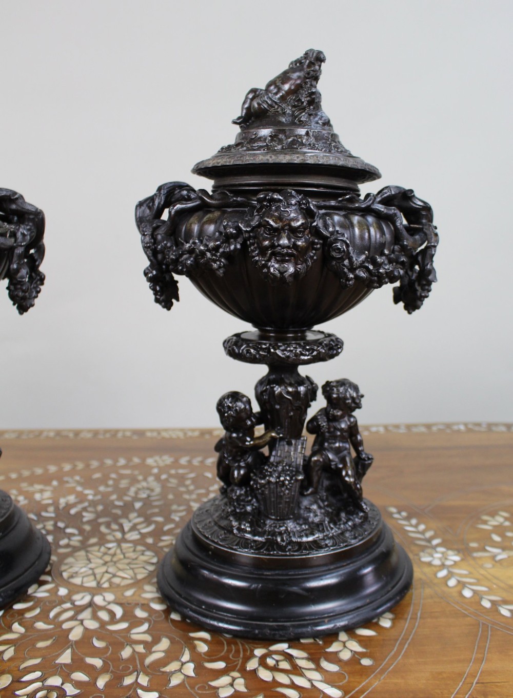 A pair of dark bronzed spelter urns with lids topped by seated putti figures, featuring Bacchic masks, relief garlands, and bases supported by sculpted cherubs. View III