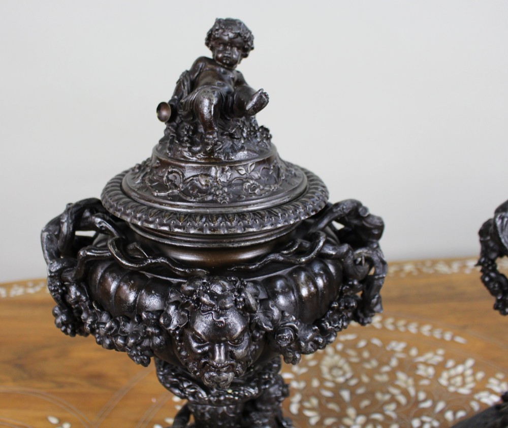 A pair of dark bronzed spelter urns with lids topped by seated putti figures, featuring Bacchic masks, relief garlands, and bases supported by sculpted cherubs. View IV