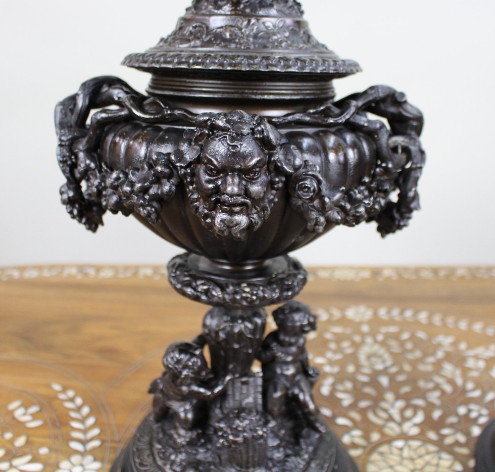 A pair of dark bronzed spelter urns with lids topped by seated putti figures, featuring Bacchic masks, relief garlands, and bases supported by sculpted cherubs. View V