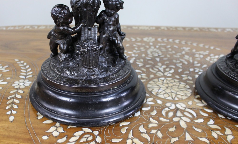 A pair of dark bronzed spelter urns with lids topped by seated putti figures, featuring Bacchic masks, relief garlands, and bases supported by sculpted cherubs. View VI