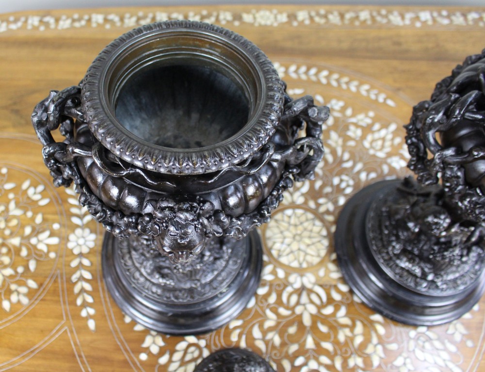 A pair of dark bronzed spelter urns with lids topped by seated putti figures, featuring Bacchic masks, relief garlands, and bases supported by sculpted cherubs. View VII