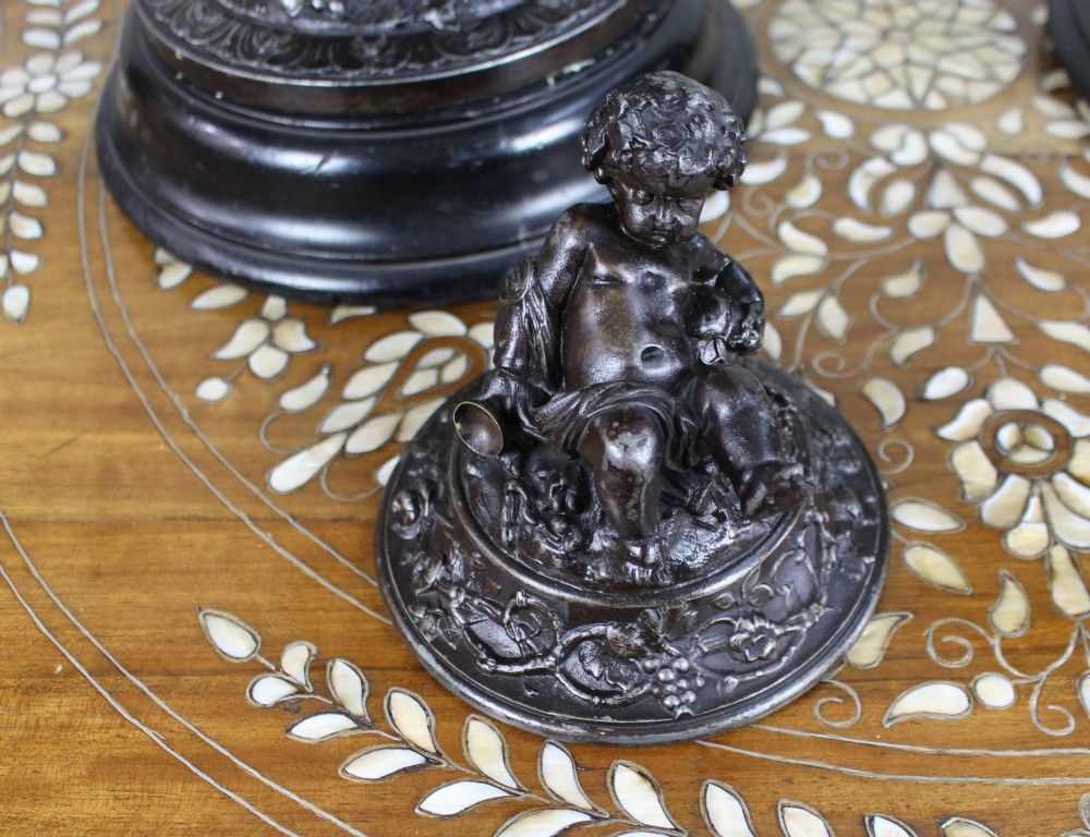 A pair of dark bronzed spelter urns with lids topped by seated putti figures, featuring Bacchic masks, relief garlands, and bases supported by sculpted cherubs. View VIII