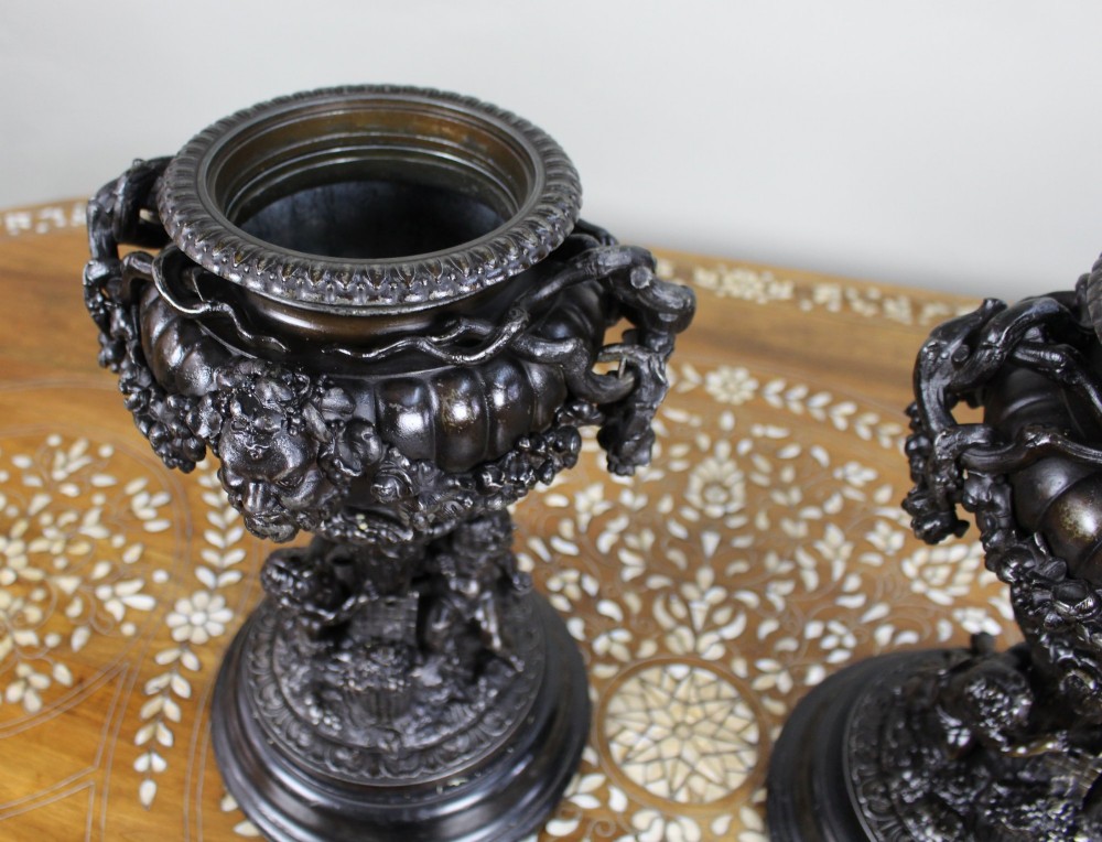A pair of dark bronzed spelter urns with lids topped by seated putti figures, featuring Bacchic masks, relief garlands, and bases supported by sculpted cherubs. View IX