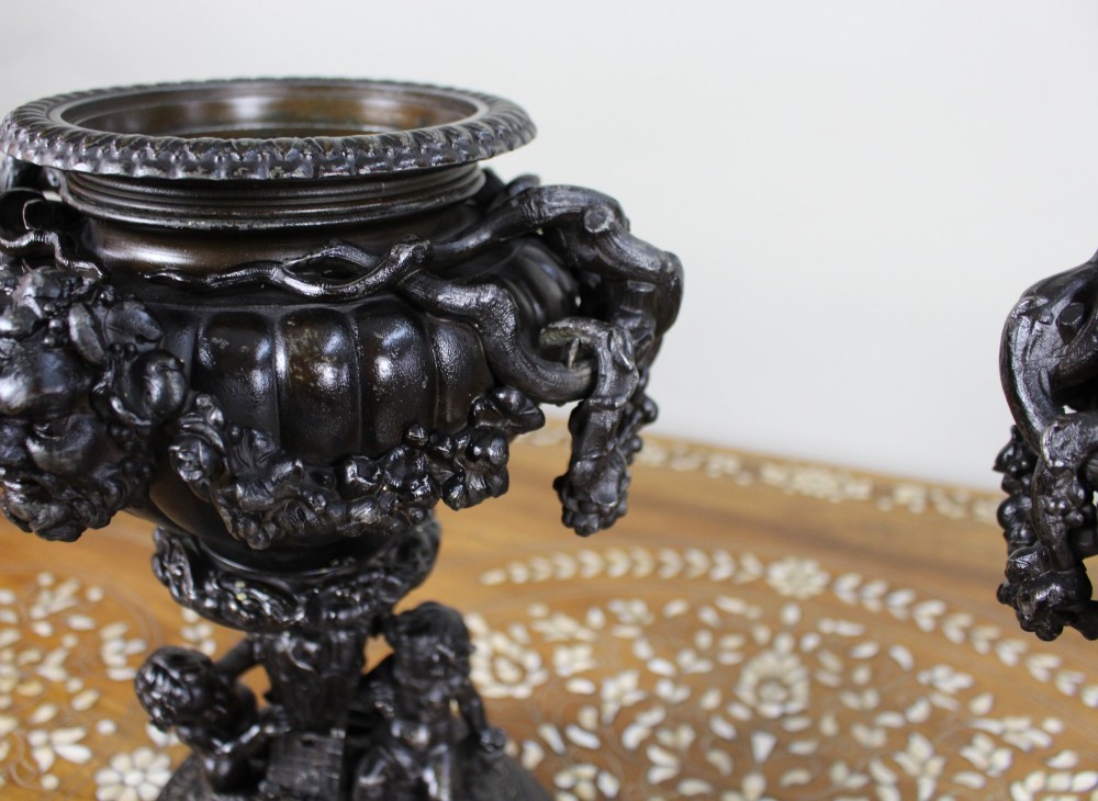 A pair of dark bronzed spelter urns with lids topped by seated putti figures, featuring Bacchic masks, relief garlands, and bases supported by sculpted cherubs. View X