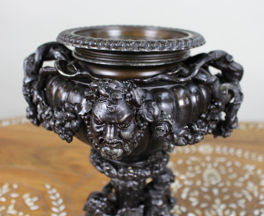 A pair of dark bronzed spelter urns with lids topped by seated putti figures, featuring Bacchic masks, relief garlands, and bases supported by sculpted cherubs. View XII