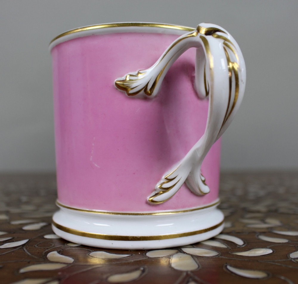 A mid-19th century cylindrical porcelain tankard featuring a Rose Pompadour pink ground, a gilded rococo scroll cartouche with hand-painted flowers, and a gilded handle. View II