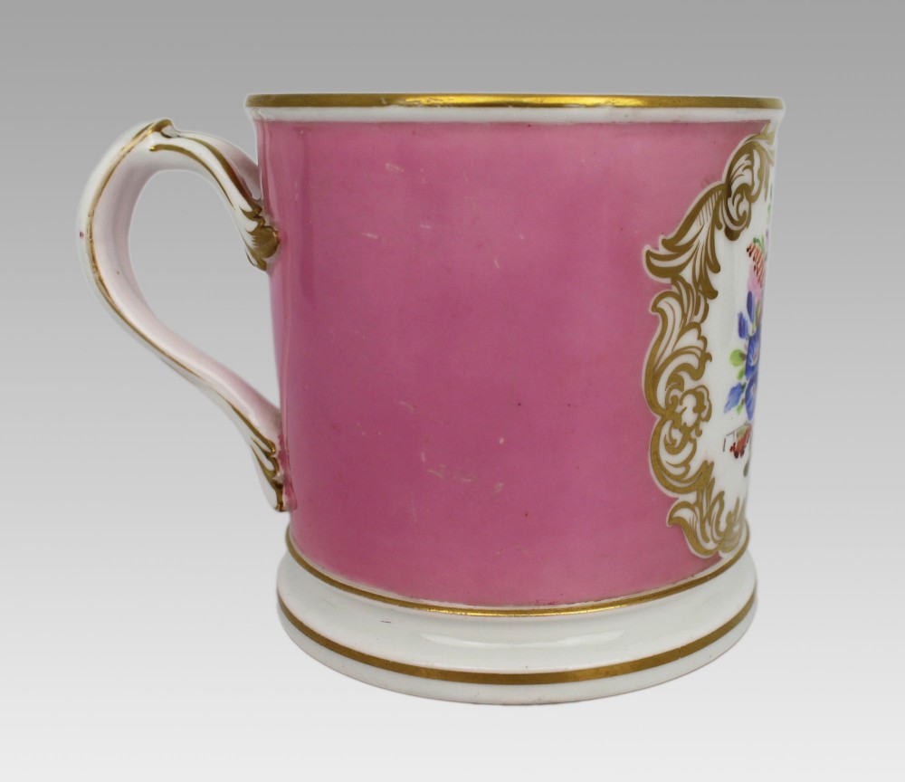 A mid-19th century cylindrical porcelain tankard featuring a Rose Pompadour pink ground, a gilded rococo scroll cartouche with hand-painted flowers, and a gilded handle. View VI