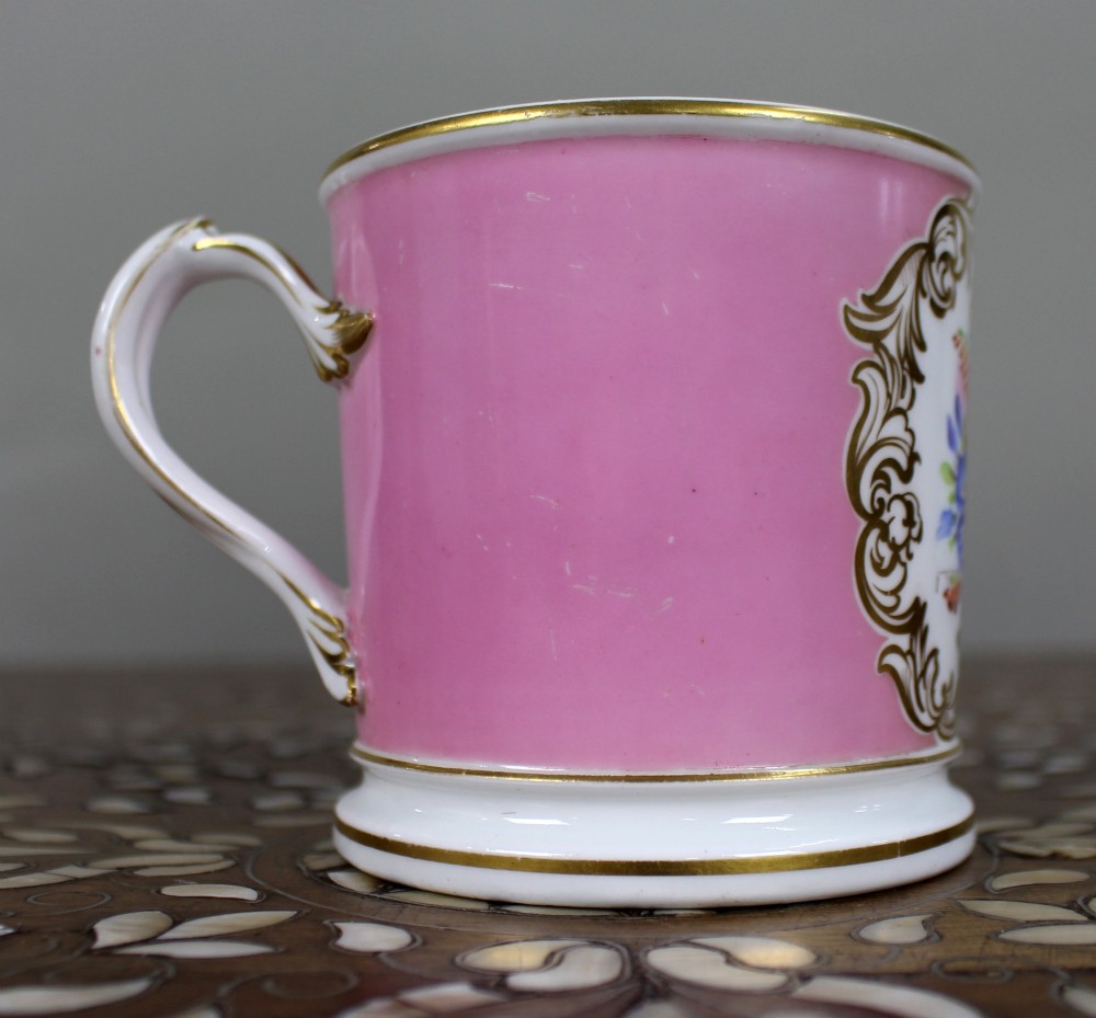 A mid-19th century cylindrical porcelain tankard featuring a Rose Pompadour pink ground, a gilded rococo scroll cartouche with hand-painted flowers, and a gilded handle. View X