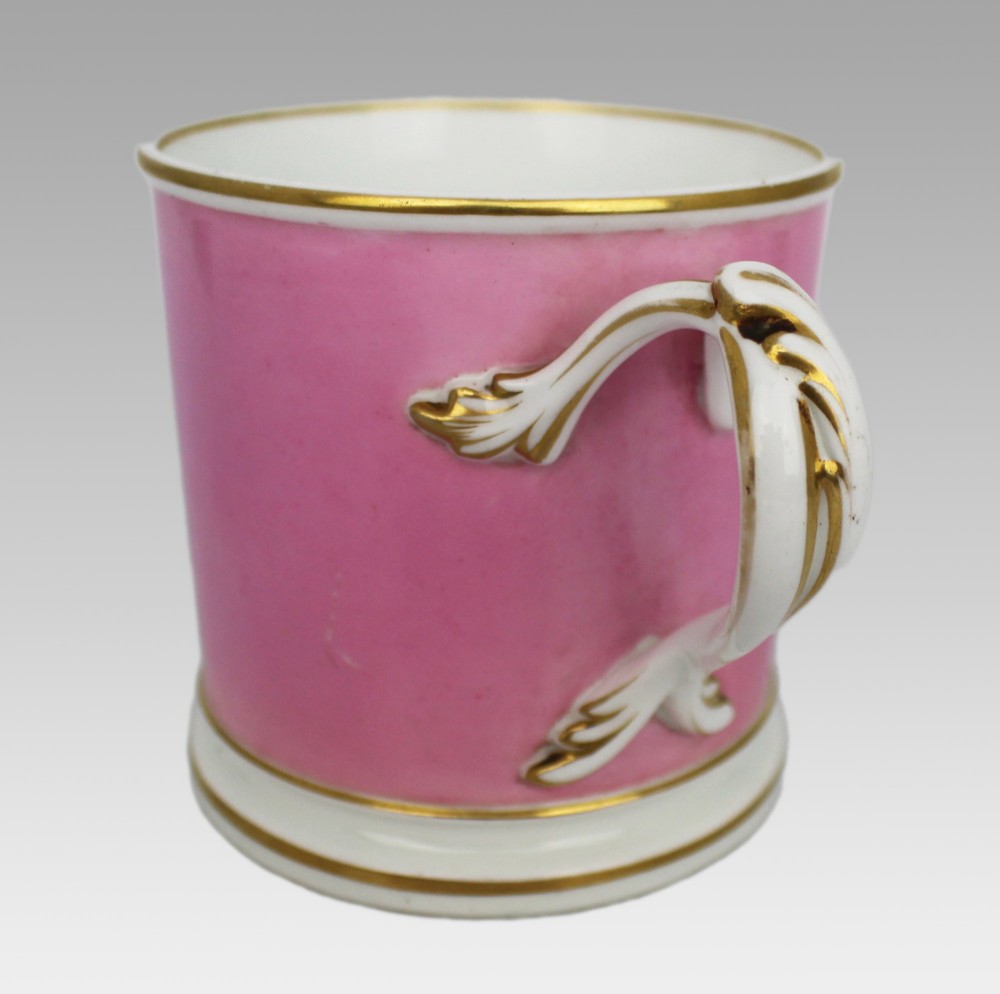 A mid-19th century cylindrical porcelain tankard featuring a Rose Pompadour pink ground, a gilded rococo scroll cartouche with hand-painted flowers, and a gilded handle. View XIII