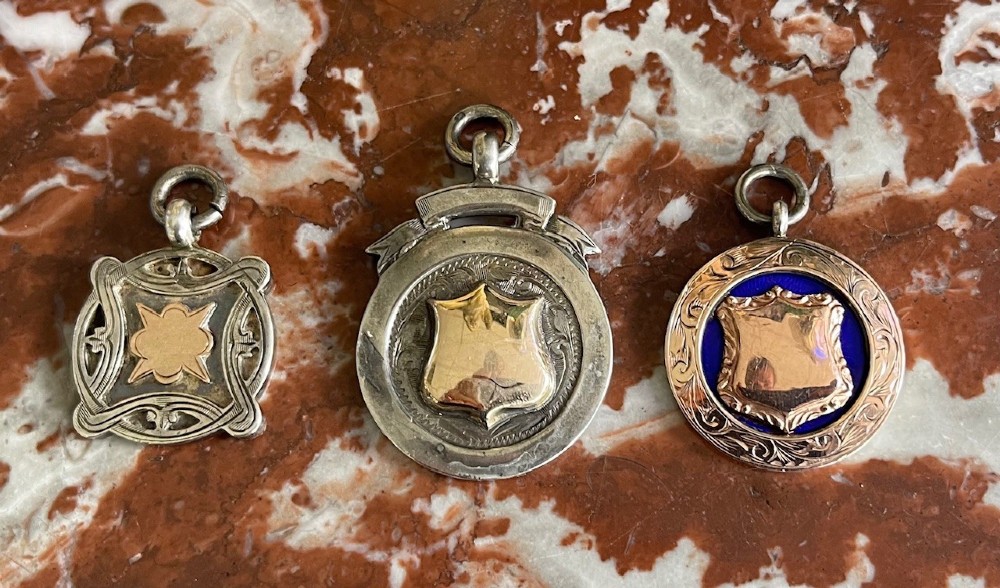 Three antique hallmarked silver watch fobs featuring central gold shield cartouches, pierced details, and blue enamel ornamentation.