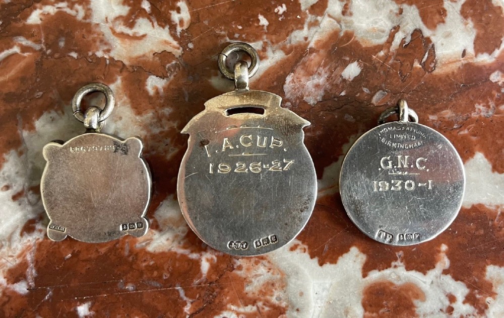 Three antique hallmarked silver watch fobs featuring central gold shield cartouches, pierced details, and blue enamel ornamentation. View II