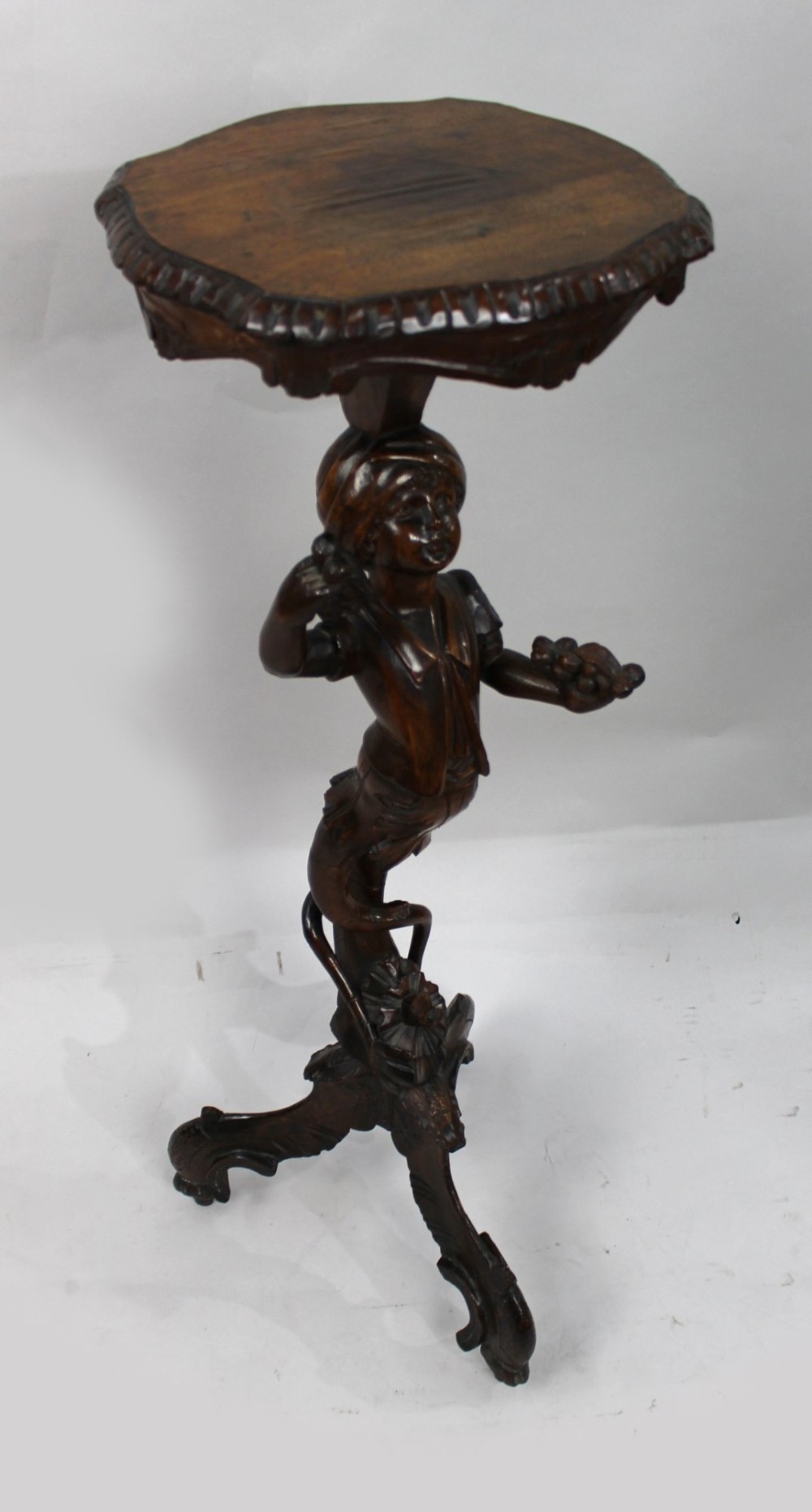 An antique dark carved wood figural pedestal featuring a central figure holding grapes supported on a tripod base with a circular scalloped top. View II