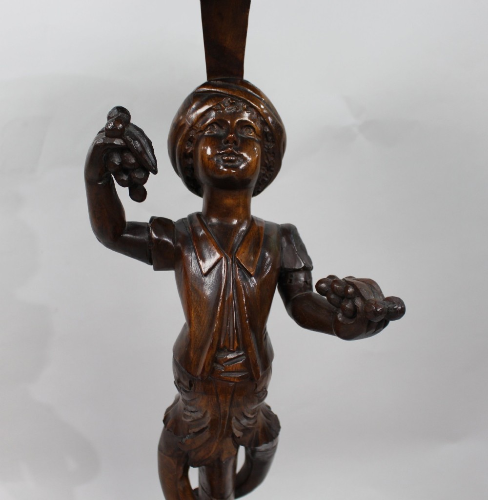 An antique dark carved wood figural pedestal featuring a central figure holding grapes supported on a tripod base with a circular scalloped top. View V