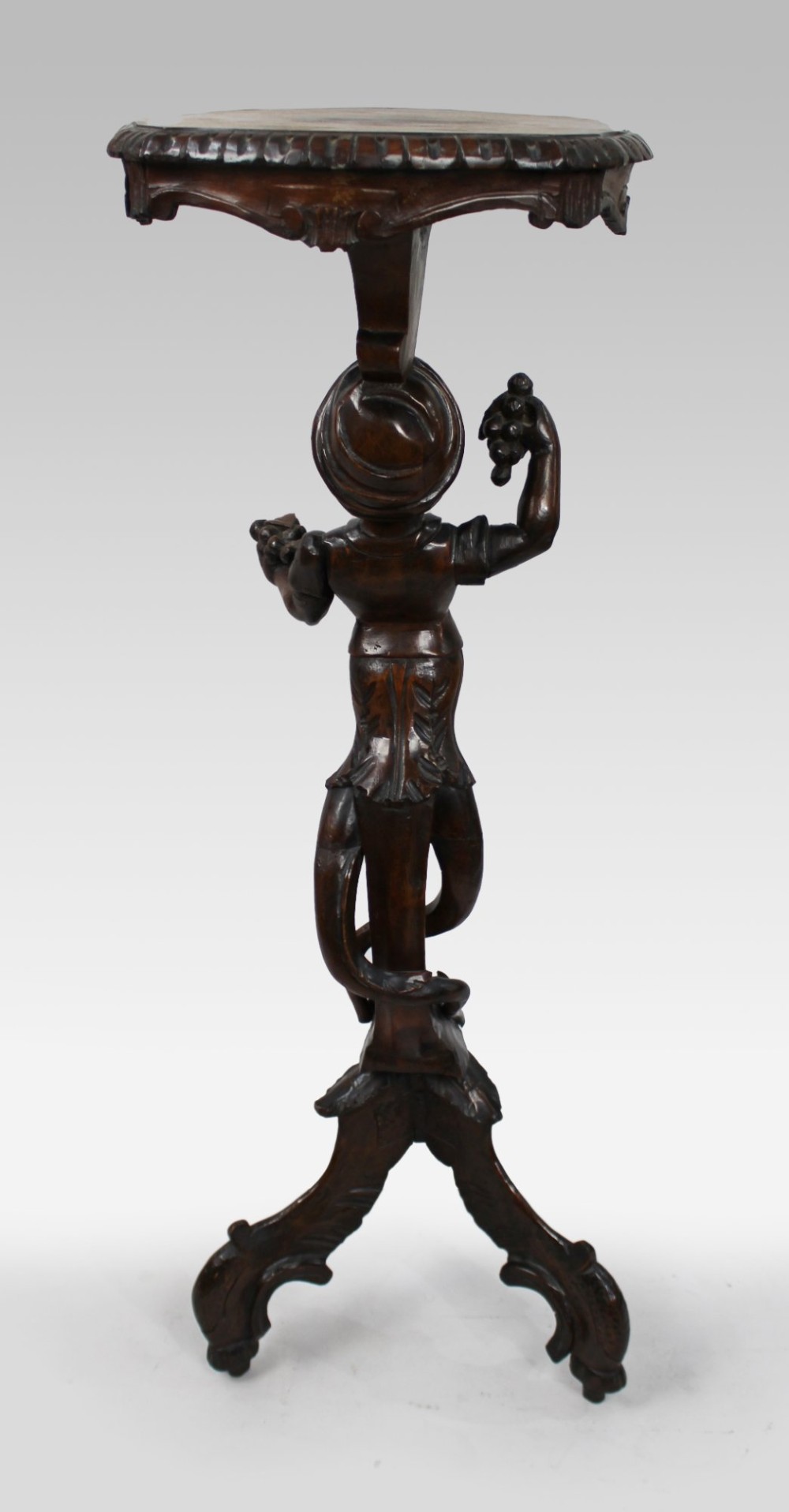 An antique dark carved wood figural pedestal featuring a central figure holding grapes supported on a tripod base with a circular scalloped top. View VIII