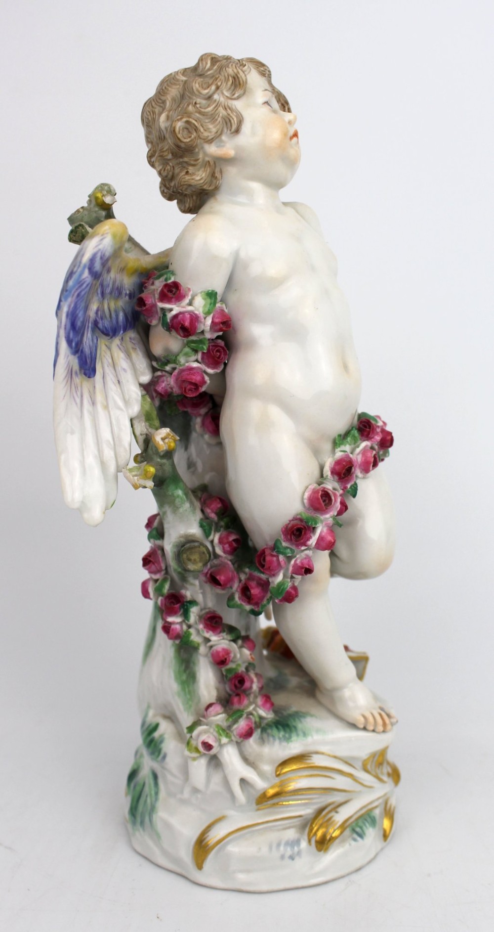 A Meissen hard-paste porcelain figurine depicting a winged cupid bound with a pink rose garland, standing on a gilded scrollwork base. View II