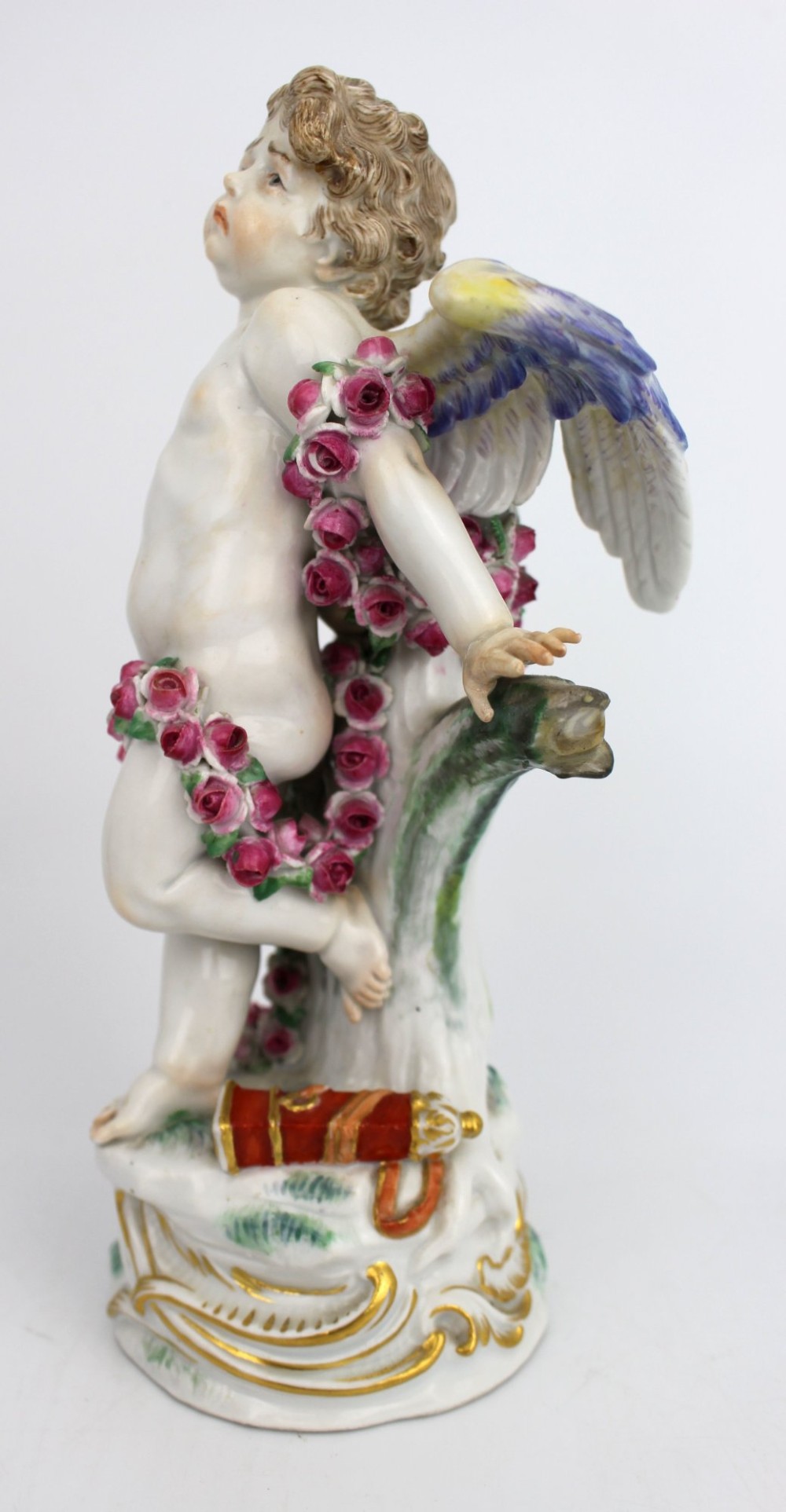 A Meissen hard-paste porcelain figurine depicting a winged cupid bound with a pink rose garland, standing on a gilded scrollwork base. View VI