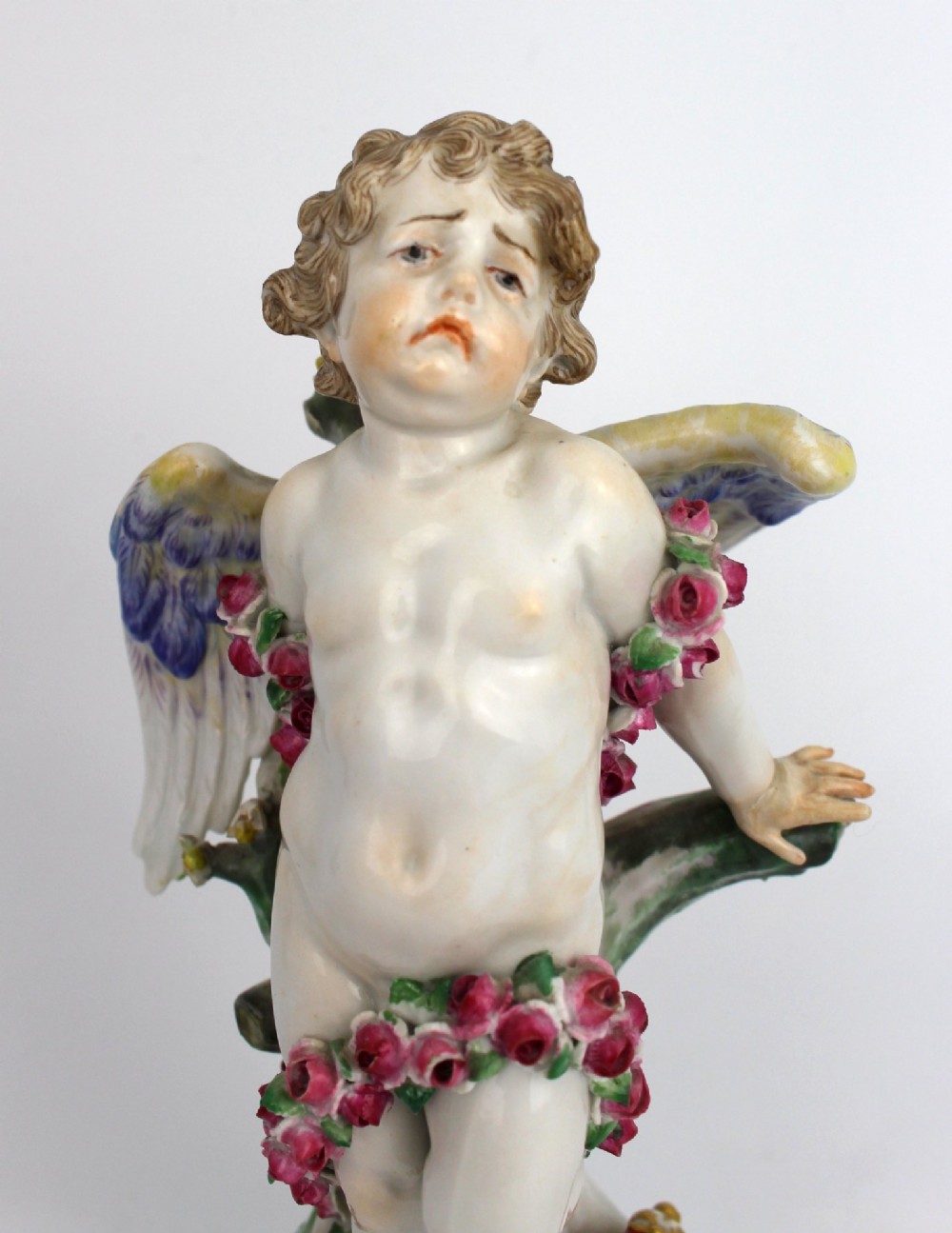 A Meissen hard-paste porcelain figurine depicting a winged cupid bound with a pink rose garland, standing on a gilded scrollwork base. View VIII