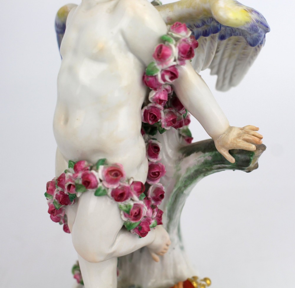 A Meissen hard-paste porcelain figurine depicting a winged cupid bound with a pink rose garland, standing on a gilded scrollwork base. View IX