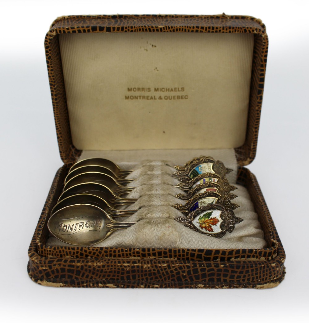 A cased set of six sterling silver demitasse spoons featuring colourful enamelled crest finials set inside a textured brown presentation box.