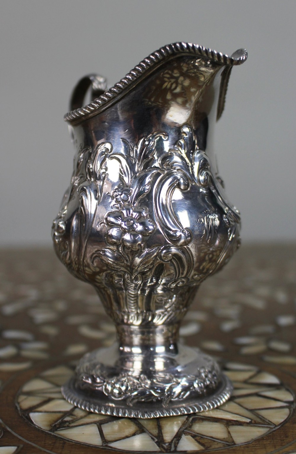 A Georgian sterling silver footed cream jug featuring floral repousse decoration, a gadrooned rim, and a curved scroll handle. View II