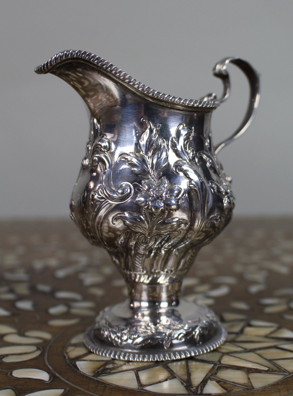 A Georgian sterling silver footed cream jug featuring floral repousse decoration, a gadrooned rim, and a curved scroll handle. View III