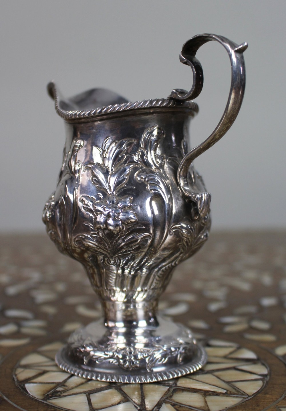 A Georgian sterling silver footed cream jug featuring floral repousse decoration, a gadrooned rim, and a curved scroll handle. View IV