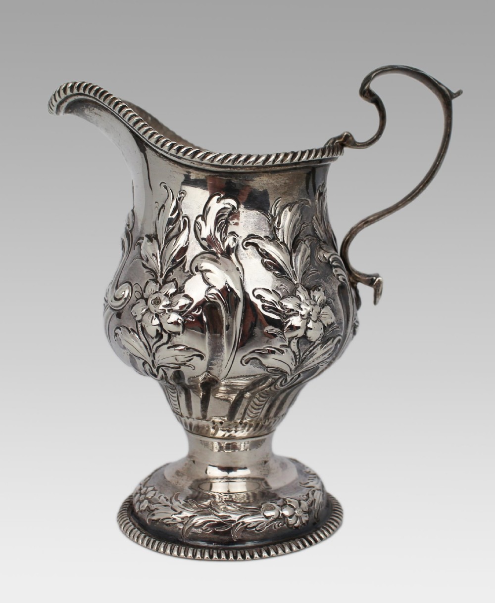 A Georgian sterling silver footed cream jug featuring floral repousse decoration, a gadrooned rim, and a curved scroll handle. View V