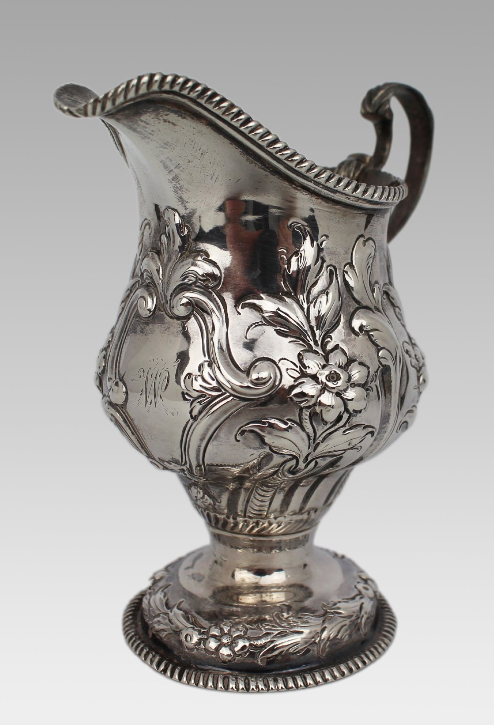 A Georgian sterling silver footed cream jug featuring floral repousse decoration, a gadrooned rim, and a curved scroll handle. View VI