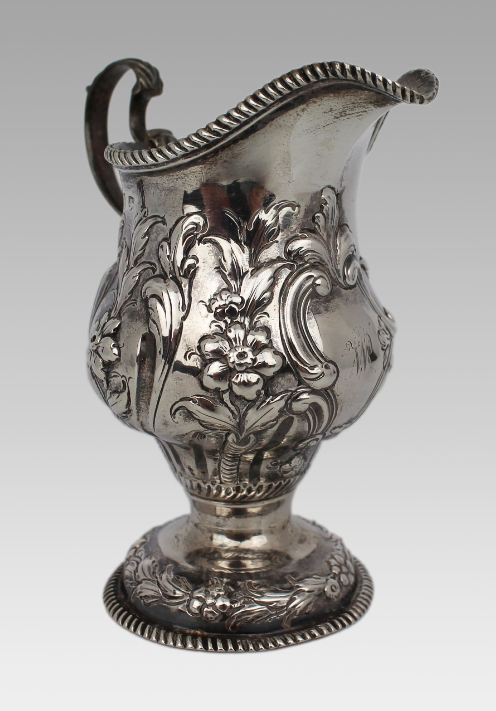 A Georgian sterling silver footed cream jug featuring floral repousse decoration, a gadrooned rim, and a curved scroll handle. View VII