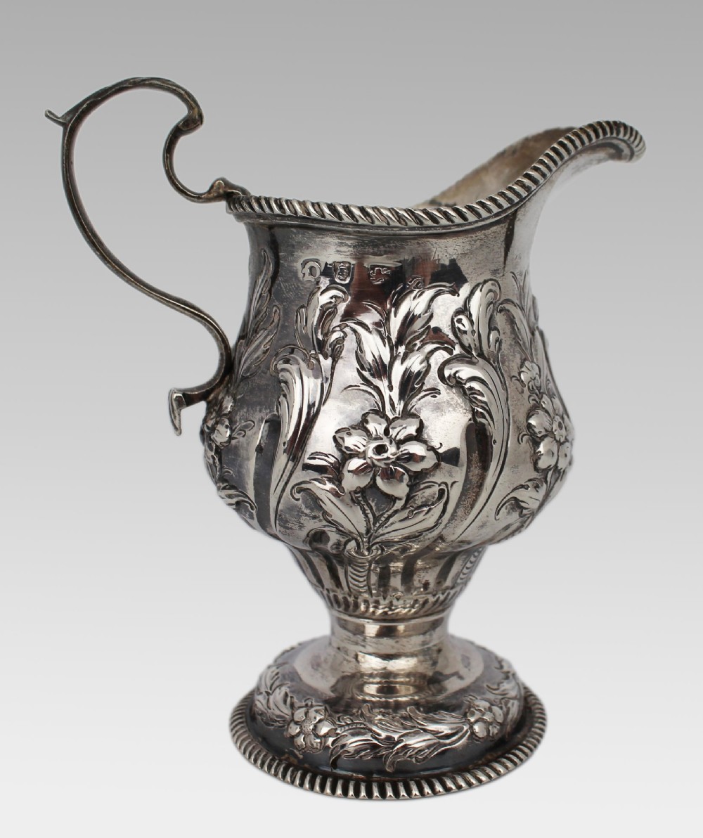 A Georgian sterling silver footed cream jug featuring floral repousse decoration, a gadrooned rim, and a curved scroll handle. View VIII