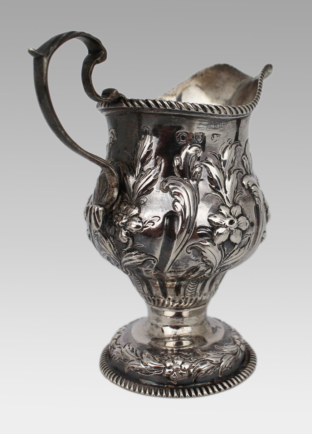 A Georgian sterling silver footed cream jug featuring floral repousse decoration, a gadrooned rim, and a curved scroll handle. View IX