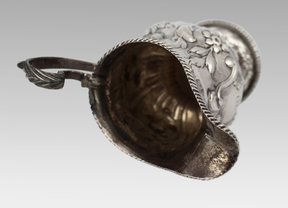 A Georgian sterling silver footed cream jug featuring floral repousse decoration, a gadrooned rim, and a curved scroll handle. View X