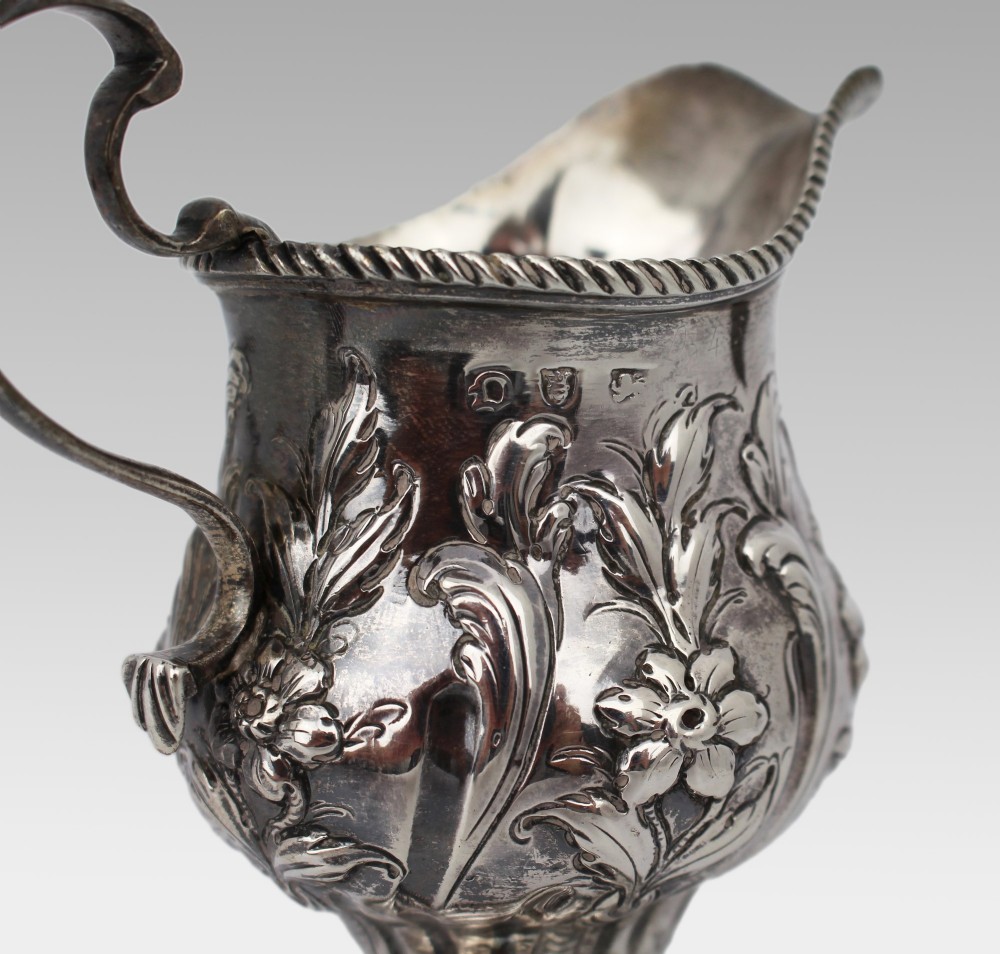 A Georgian sterling silver footed cream jug featuring floral repousse decoration, a gadrooned rim, and a curved scroll handle. View XI