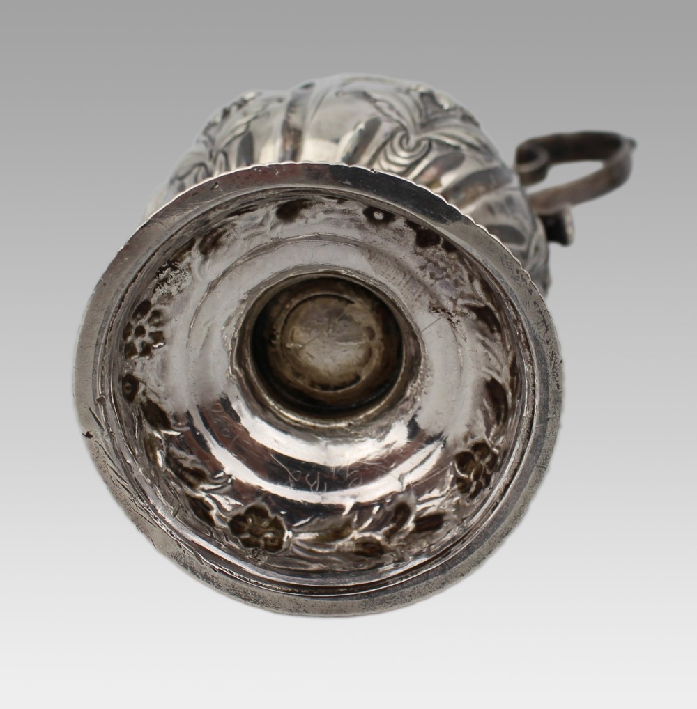 A Georgian sterling silver footed cream jug featuring floral repousse decoration, a gadrooned rim, and a curved scroll handle. View XII