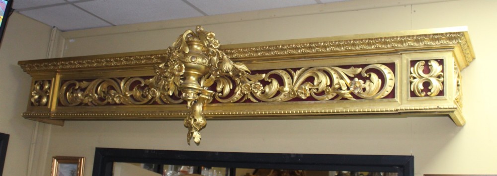 A Regency gilded wooden window pelmet featuring carved acanthus scrolls, floral swags, and a central urn motif set against a dark red fabric backing.