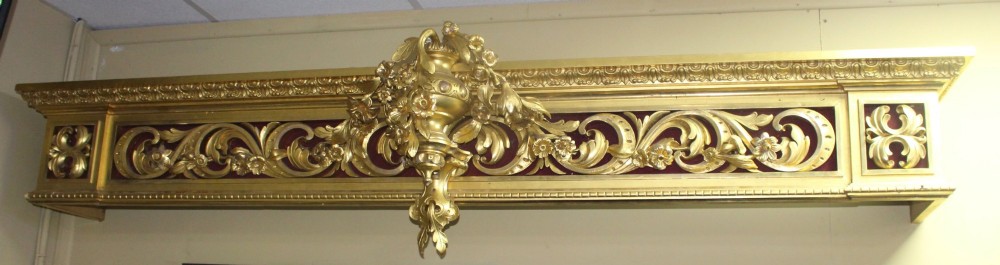 A Regency gilded wooden window pelmet featuring carved acanthus scrolls, floral swags, and a central urn motif set against a dark red fabric backing. View II