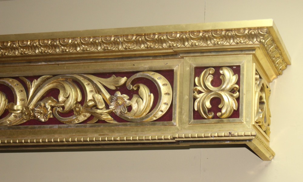 A Regency gilded wooden window pelmet featuring carved acanthus scrolls, floral swags, and a central urn motif set against a dark red fabric backing. View VI