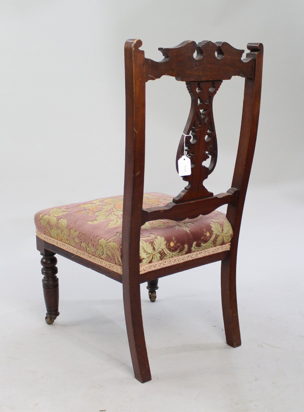 An Edwardian mahogany nursing chair featuring a carved pierced backrest, low padded floral tapestry seat, and turned front legs with castors. View III
