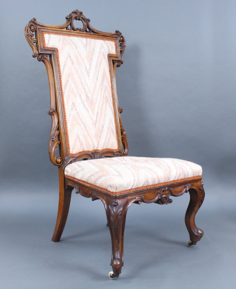A Victorian carved walnut occasional chair featuring a tall upholstered backrest with patterned beige fabric, scroll-carved cresting, cabriole legs, and ceramic castors.