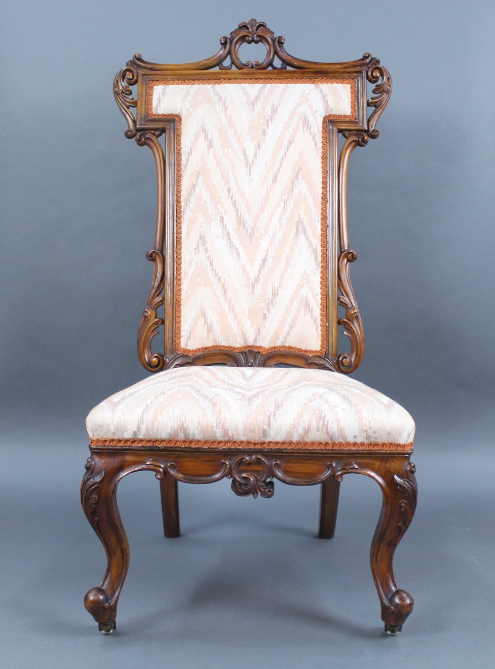 A Victorian carved walnut occasional chair featuring a tall upholstered backrest with patterned beige fabric, scroll-carved cresting, cabriole legs, and ceramic castors. View II
