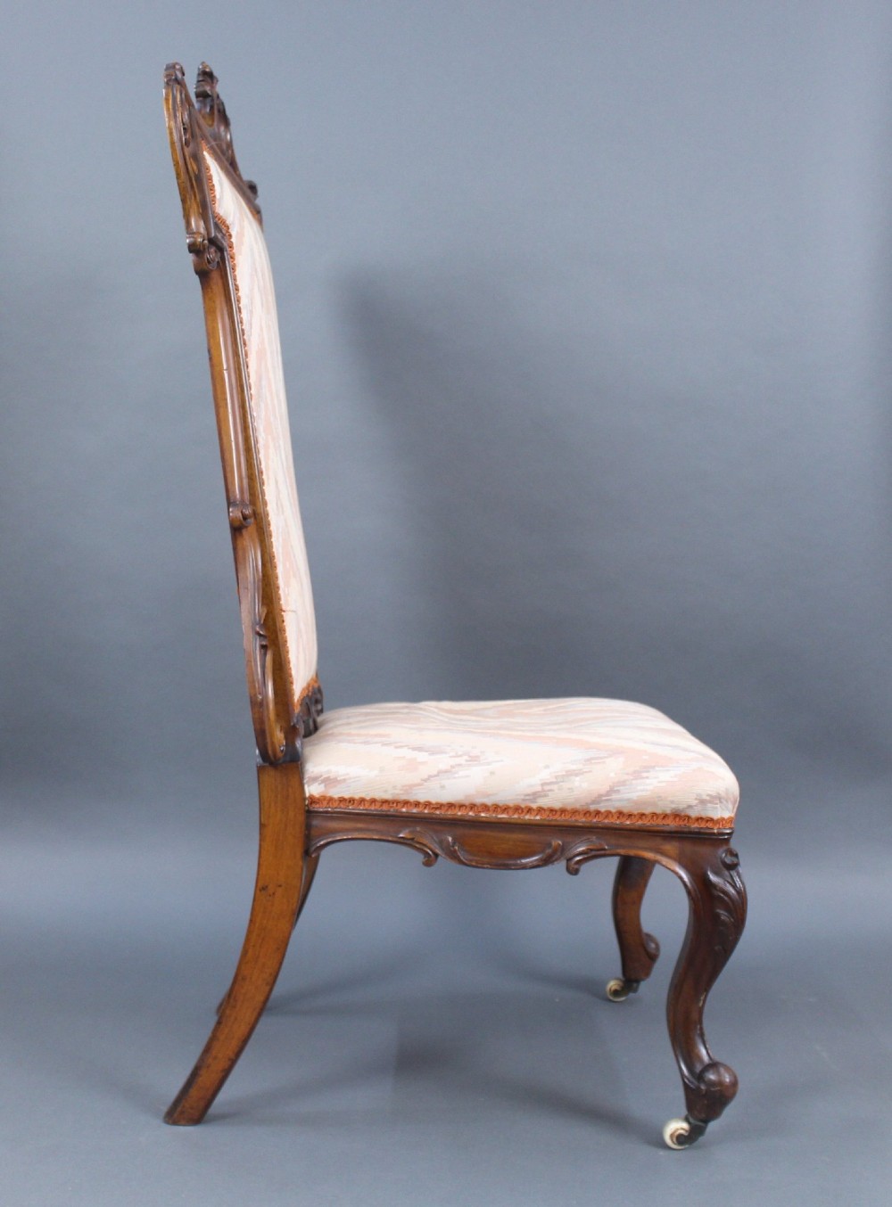 A Victorian carved walnut occasional chair featuring a tall upholstered backrest with patterned beige fabric, scroll-carved cresting, cabriole legs, and ceramic castors. View III