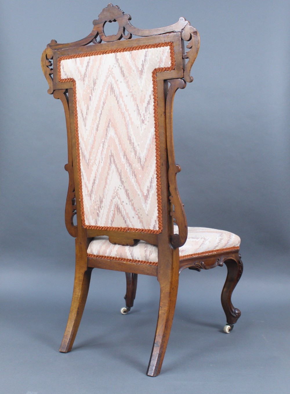 A Victorian carved walnut occasional chair featuring a tall upholstered backrest with patterned beige fabric, scroll-carved cresting, cabriole legs, and ceramic castors. View IV