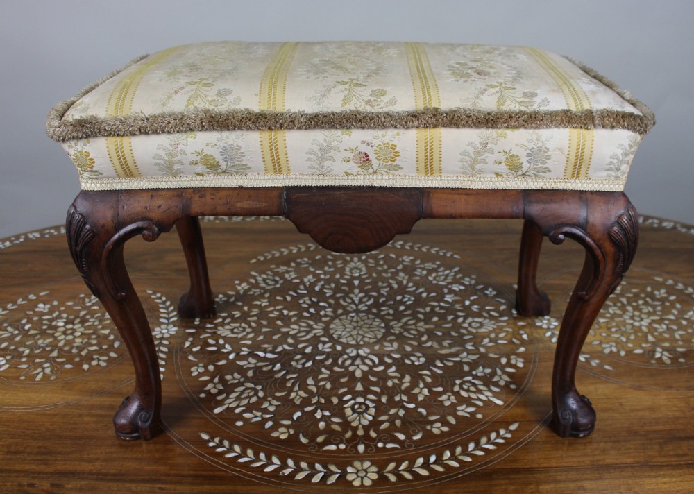 A low walnut footstool featuring carved cabriole legs and a cream and yellow floral striped fabric upholstered seat with tassel trim.