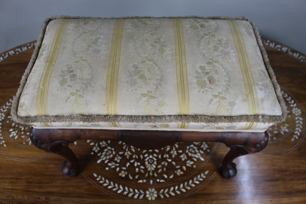 A low walnut footstool featuring carved cabriole legs and a cream and yellow floral striped fabric upholstered seat with tassel trim. View II